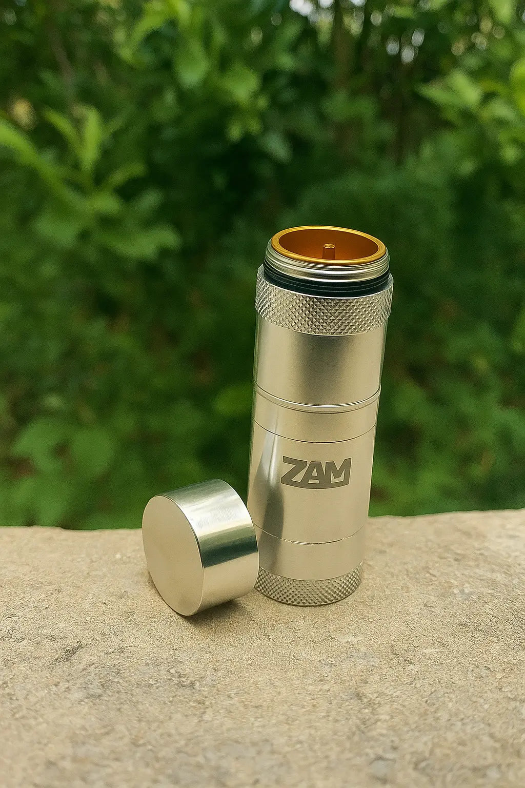 Metallic cylindrical object with 'ZAM' branding on a stone surface with green foliage in the background